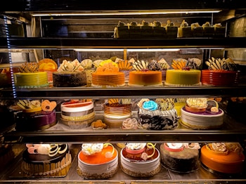 An assortment of colorful and intricately decorated cakes are displayed on shelves in a bakery. The cakes vary in flavor and design, with layers of frosting, fruit, chocolate, and decorative toppings prominently featured. Some of the cakes are round while others have more unique shapes, all presented in an enticing and artistic manner.