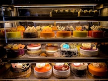 An assortment of colorful and intricately decorated cakes are displayed on shelves in a bakery. The cakes vary in flavor and design, with layers of frosting, fruit, chocolate, and decorative toppings prominently featured. Some of the cakes are round while others have more unique shapes, all presented in an enticing and artistic manner.