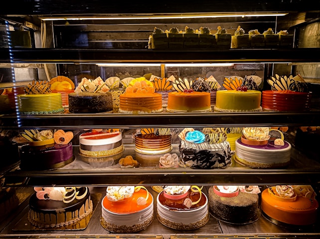 An assortment of colorful and intricately decorated cakes are displayed on shelves in a bakery. The cakes vary in flavor and design, with layers of frosting, fruit, chocolate, and decorative toppings prominently featured. Some of the cakes are round while others have more unique shapes, all presented in an enticing and artistic manner.