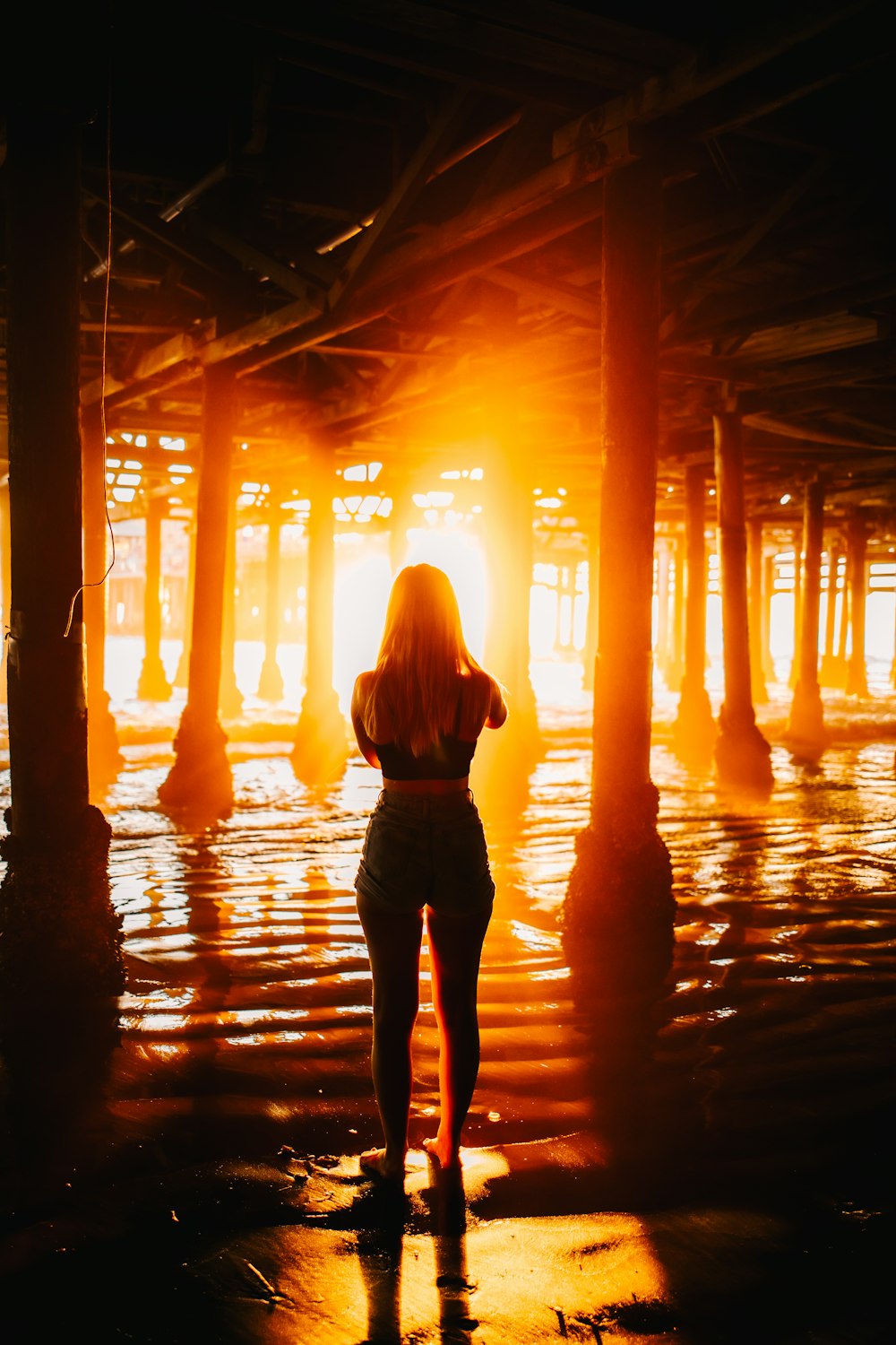 a woman standing under a pier at sunset