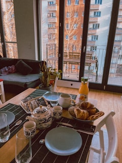 A cozy dining setup is arranged on a wooden table, featuring blue plates, black placemats, and an ornate white and blue teapot with matching cups. A wicker basket contains a variety of breads and pastries. There is also a bottle of orange juice on the table. Large glass doors in the background open to a view of neighboring buildings, with sunlight filtering through.