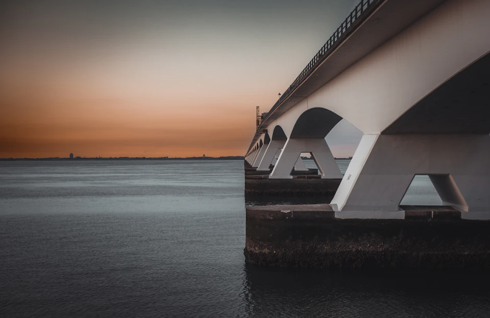White concrete bridge with four arches spans across calm water under orange-pink sky
