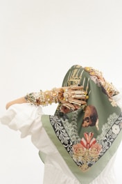 A person wearing a decorative green headscarf adorned with intricate floral and nature-inspired patterns in shades of red, white, and gold. The person's arms are covered with numerous layered bracelets made of pearls and gemstones, adding an ornate and eclectic touch to the ensemble. The clothing appears to be white and flowing.