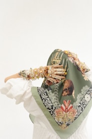 A person wearing a decorative green headscarf adorned with intricate floral and nature-inspired patterns in shades of red, white, and gold. The person's arms are covered with numerous layered bracelets made of pearls and gemstones, adding an ornate and eclectic touch to the ensemble. The clothing appears to be white and flowing.