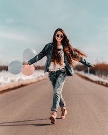 A person with long brown hair, wearing sunglasses and a denim outfit, is joyfully walking down a deserted road. They are holding colorful balloons with a big smile, creating a sense of freedom and happiness.
