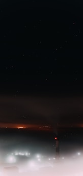 A tall smokestack emits smoke under a dark night sky. The atmosphere is hazy with a mix of fog and industrial emissions. Distant lights glow faintly, creating an eerie and industrial scene. The night is starless, and the smoke appears to blend into the darkness.