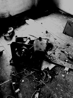A dilapidated room with a broken appliance lying on the wooden floor surrounded by debris, wires, and dust. The room exhibits signs of neglect with peeling paint and dirt.