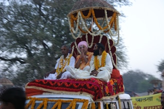 A group of three individuals sitting on a decorated platform atop a vehicle, adorned with vibrant fabrics and festive garlands. One of them is wearing a pink turban, and all are draped in floral garlands. The background consists of blurred trees, suggesting movement.