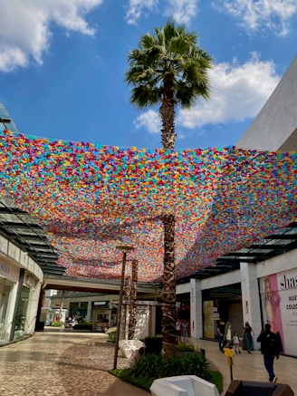 A vibrant outdoor shopping area with a decorative canopy made of colorful, suspended materials resembling hundreds of small tiles. The canopy creates an artistic effect, casting brilliant patterns of light on the ground below. Tall palm trees punctuate the space, and shoppers can be seen strolling past various storefronts.