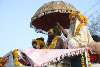 Several people adorned with yellow and orange garlands are sitting on top of a vehicle. An ornate umbrella is held above them, providing shade. One individual is wearing a white robe and has a long beard. Bright sunlight illuminates the scene.