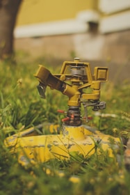 A close-up view of an old, yellow garden sprinkler atop green grass, showing signs of wear and rust. The blurred background includes a tree trunk and part of a yellow and white building.