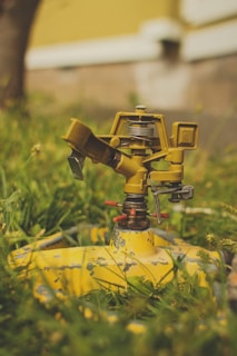 A close-up view of an old, yellow garden sprinkler atop green grass, showing signs of wear and rust. The blurred background includes a tree trunk and part of a yellow and white building.