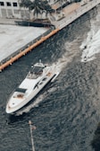 A large yacht navigates through a canal bordered by a dock on one side. Two jetskis speed alongside, creating white trails in the blue-green water. Palm trees and modern buildings line the background, suggesting a coastal or urban waterfront setting. An American flag is visible in the lower foreground.
