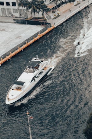 A large yacht navigates through a canal bordered by a dock on one side. Two jetskis speed alongside, creating white trails in the blue-green water. Palm trees and modern buildings line the background, suggesting a coastal or urban waterfront setting. An American flag is visible in the lower foreground.