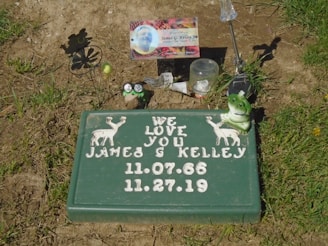 A memorial site features a green plaque with white text and illustrations of two deer. It reads 'We Love You James G Kelley' along with dates '11.07.66' and '11.27.19'. The area is adorned with small items including a frog ornament, a spinning flower silhouette, a small picture, and a few other mementos resting on the ground and grass.