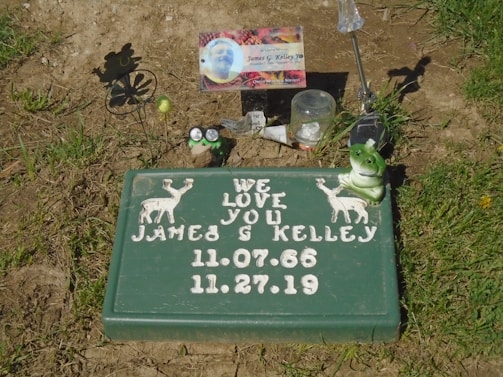 A memorial site features a green plaque with white text and illustrations of two deer. It reads 'We Love You James G Kelley' along with dates '11.07.66' and '11.27.19'. The area is adorned with small items including a frog ornament, a spinning flower silhouette, a small picture, and a few other mementos resting on the ground and grass.