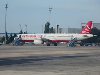 A passenger airplane with a red and white color scheme is stationed on an airport runway. The aircraft has its name and logo prominently displayed on the fuselage and tail. Portable staircases are positioned at both the front and rear doors of the airplane. The surroundings include airport infrastructure such as buildings, poles, and service vehicles.