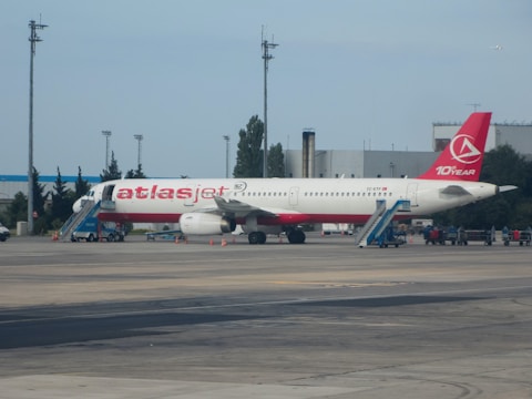 A passenger airplane with a red and white color scheme is stationed on an airport runway. The aircraft has its name and logo prominently displayed on the fuselage and tail. Portable staircases are positioned at both the front and rear doors of the airplane. The surroundings include airport infrastructure such as buildings, poles, and service vehicles.