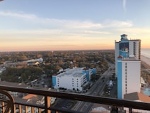 A view from a high vantage point overlooks a coastal cityscape. In the foreground, a balcony railing frames the scene. Several tall buildings with blue accents and red roofs are prominently visible, alongside a road that leads towards a horizon filled with dense, green foliage. The sky is partly cloudy, with hues of orange and pink due to the setting sun, blending into a serene blue.