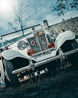 A vintage white car with classic round headlights and a chrome grille is parked on a slightly grassy surface. The angle of the photo is low, emphasizing the car's imposing front view. The car bears two red medallions and a Union Jack emblem. The background features a cloudy sky and some blurred foliage.