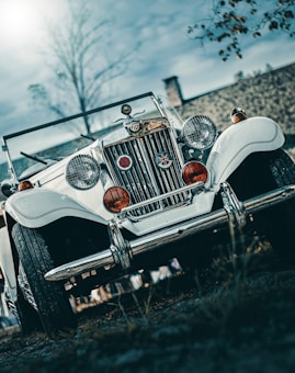 A vintage white car with classic round headlights and a chrome grille is parked on a slightly grassy surface. The angle of the photo is low, emphasizing the car's imposing front view. The car bears two red medallions and a Union Jack emblem. The background features a cloudy sky and some blurred foliage.