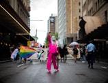 A person dressed in a bright pink outfit with long hair and a mask stands confidently on a city street, surrounded by a diverse crowd of protesters carrying colorful banners and signs advocating for equality and against discrimination. The scene is set on a cloudy day in an urban environment, with tall buildings lining the street.