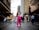 A person dressed in a bright pink outfit with long hair and a mask stands confidently on a city street, surrounded by a diverse crowd of protesters carrying colorful banners and signs advocating for equality and against discrimination. The scene is set on a cloudy day in an urban environment, with tall buildings lining the street.