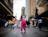A person dressed in a bright pink outfit with long hair and a mask stands confidently on a city street, surrounded by a diverse crowd of protesters carrying colorful banners and signs advocating for equality and against discrimination. The scene is set on a cloudy day in an urban environment, with tall buildings lining the street.