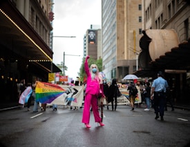 A person dressed in a bright pink outfit with long hair and a mask stands confidently on a city street, surrounded by a diverse crowd of protesters carrying colorful banners and signs advocating for equality and against discrimination. The scene is set on a cloudy day in an urban environment, with tall buildings lining the street.