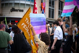 A diverse group of people is gathered in a city street, some holding up signs. One prominent sign reads 'LOVE TRANS KIDS' with a background of blue, pink, and white stripes. Other signs are partially visible. Many attendees are wearing face masks, and there is a variety of clothing styles. The background shows a building with the word 'galleries' and several flags.