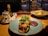 A warmly lit table setting in a restaurant features a main dish of toasted bread topped with melted cheese and microgreens, accompanied by a thick, dark sauce. Nearby, a fresh salad includes pumpkin and feta cheese, all on colorful plates. The table also holds condiments like olive oil and a pepper mill, while city lights are visible through a window in the background.