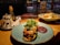 A warmly lit table setting in a restaurant features a main dish of toasted bread topped with melted cheese and microgreens, accompanied by a thick, dark sauce. Nearby, a fresh salad includes pumpkin and feta cheese, all on colorful plates. The table also holds condiments like olive oil and a pepper mill, while city lights are visible through a window in the background.