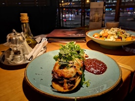 A warmly lit table setting in a restaurant features a main dish of toasted bread topped with melted cheese and microgreens, accompanied by a thick, dark sauce. Nearby, a fresh salad includes pumpkin and feta cheese, all on colorful plates. The table also holds condiments like olive oil and a pepper mill, while city lights are visible through a window in the background.