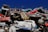 A pile of old, rusted cars stacked haphazardly on top of each other under a clear blue sky. The vehicles are in various stages of decay, with missing parts and visible damage.