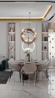A modern dining room with a long table set for six, featuring elegant minimalist chairs and a striking geometric chandelier with spherical bulbs. The room has built-in shelving units with decorative items and books, and a comfortable green sofa with a geometric rug on the side. The floor is glossy marble with a subtle pattern, adding a touch of luxury.