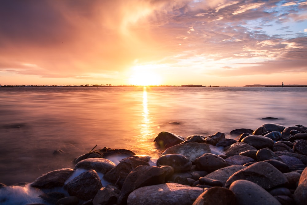 un coucher de soleil sur un plan d’eau avec des rochers au premier plan