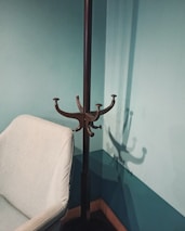 A minimalist interior corner featuring a light gray chair with a metal coat rack next to it. The wall and floor have a teal color, providing a calm and modern look. The coat rack casts a distinct shadow on the wall.