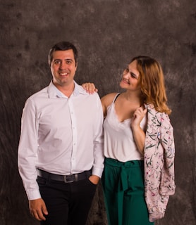 A smiling man in a white shirt stands next to a woman with shoulder-length hair who is looking at him with a smile. The woman is wearing a white camisole under a floral jacket draped over her shoulder and green pants.