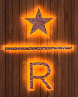 A wooden background features a prominent illuminated star above a horizontal line and the letter R. The star and letter are made from a textured metallic material, with an orange glow emanating from behind.