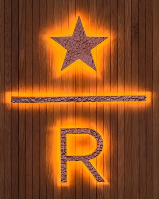 A wooden background features a prominent illuminated star above a horizontal line and the letter R. The star and letter are made from a textured metallic material, with an orange glow emanating from behind.