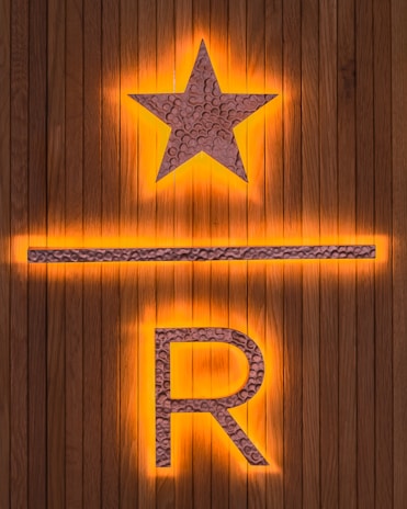 A wooden background features a prominent illuminated star above a horizontal line and the letter R. The star and letter are made from a textured metallic material, with an orange glow emanating from behind.