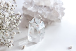 A clear quartz crystal with a silver crescent moon ring around it is surrounded by delicate white baby's breath flowers. The background consists of blurred, light-colored crystals, creating a soft and serene setting.