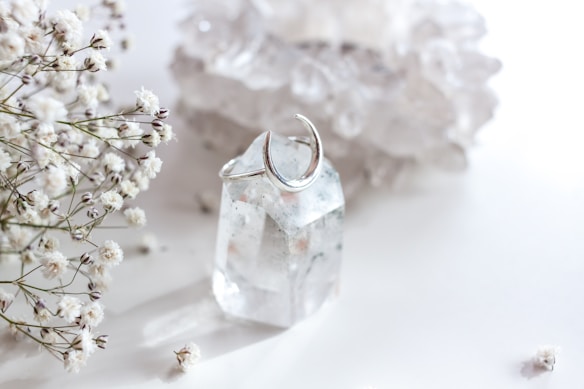A clear quartz crystal with a silver crescent moon ring around it is surrounded by delicate white baby's breath flowers. The background consists of blurred, light-colored crystals, creating a soft and serene setting.