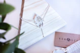 A crystal necklace with a delicate silver chain is elegantly displayed on a light wooden, pyramid-shaped stand. To the side, a business card reads 'ESMAGIC' with mystical-themed branding. A green plant is partially visible in the foreground, adding a touch of nature to the composition.