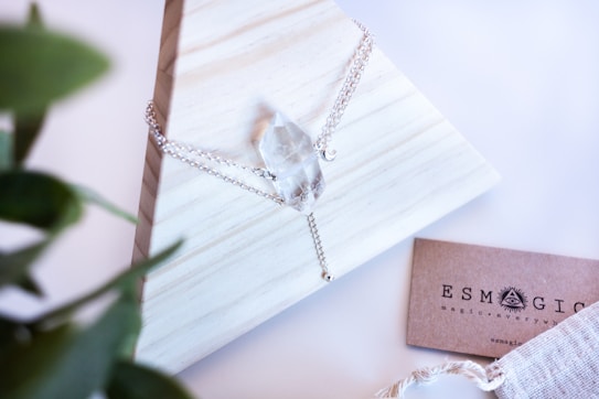 A crystal necklace with a delicate silver chain is elegantly displayed on a light wooden, pyramid-shaped stand. To the side, a business card reads 'ESMAGIC' with mystical-themed branding. A green plant is partially visible in the foreground, adding a touch of nature to the composition.