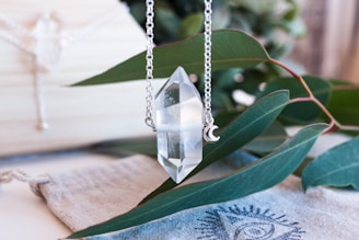 A clear crystal pendant hangs from a silver chain. The background features green leaves and a fabric piece with a printed eye design. The setting is airy and natural, with soft focus elements that suggest a serene environment.