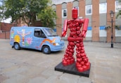 A sculpture of a human figure made from red fire extinguishers stands on a wooden platform in an urban setting. Nearby, a blue van features colorful mural artwork with designs such as a totem pole, dragons, and 'Ancestors Art' written on the side. The background is a brick building with windows and a tree, giving a vibrant and artistic urban feel.