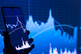 A hand holds a smartphone displaying a financial chart with candlestick patterns. In the background, a large, similar financial graph is visible on a screen, featuring fluctuating lines and data points.