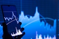 A hand holds a smartphone displaying a financial chart with candlestick patterns. In the background, a large, similar financial graph is visible on a screen, featuring fluctuating lines and data points.