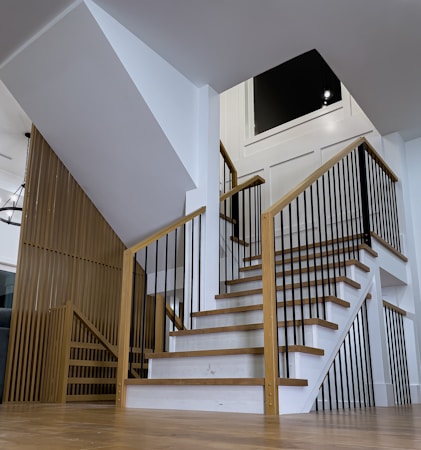 A modern staircase with wooden steps and black metal railings, leading up from a wooden floor. White walls and an open layout create a minimalist and spacious atmosphere.